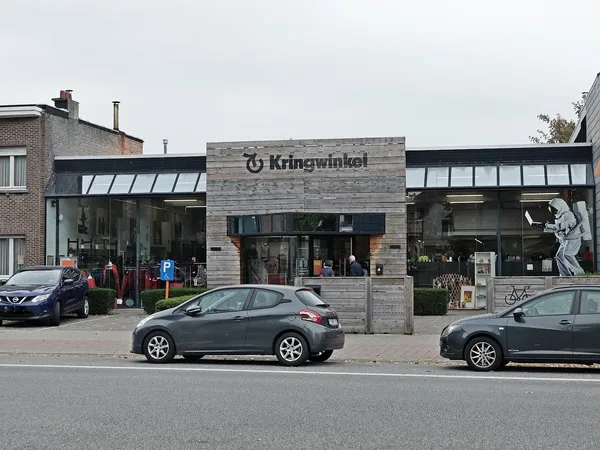 Gevel van kringloopwinkel met stenen voorgevel en grote ramen, parkeerplaats ervoor met auto's