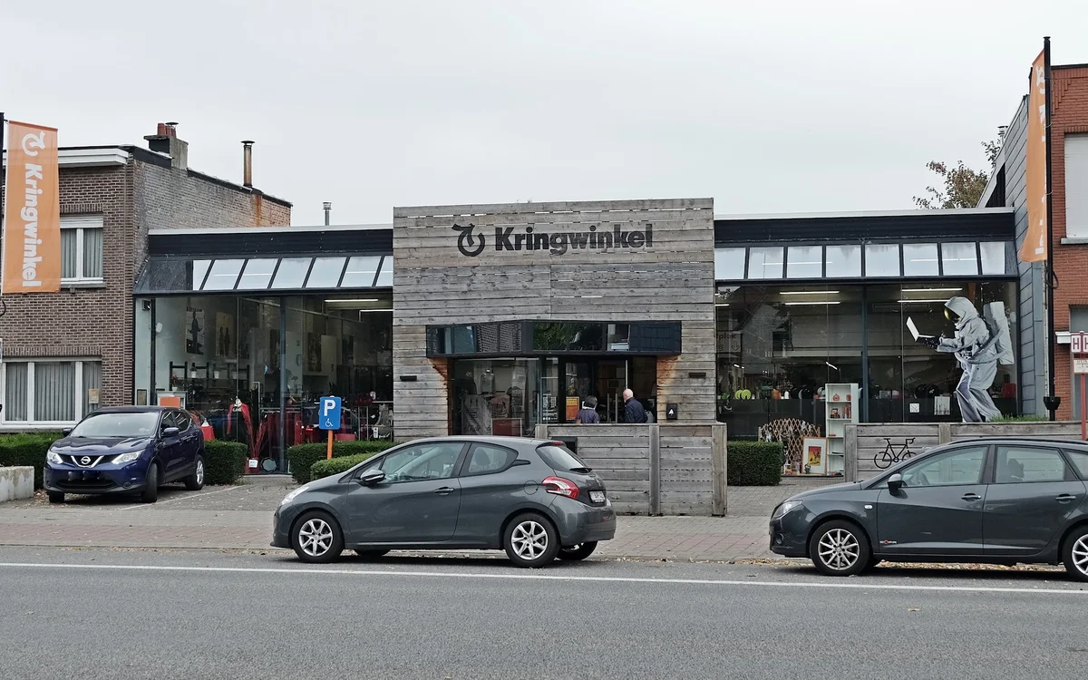 Gevel van kringloopwinkel met stenen voorgevel en grote ramen, parkeerplaats ervoor met auto's