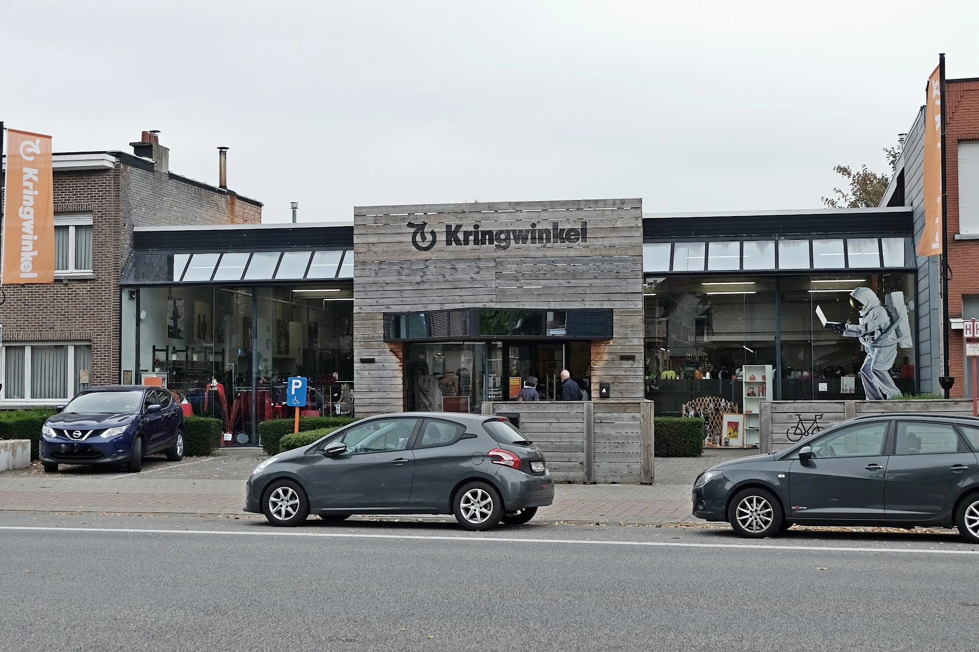 Gevel van kringloopwinkel met stenen voorgevel en grote ramen, parkeerplaats ervoor met auto's