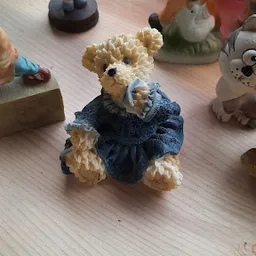 Knuffelbeer van beige pluche in blauw jurkje met strik, staand op houten vloer.