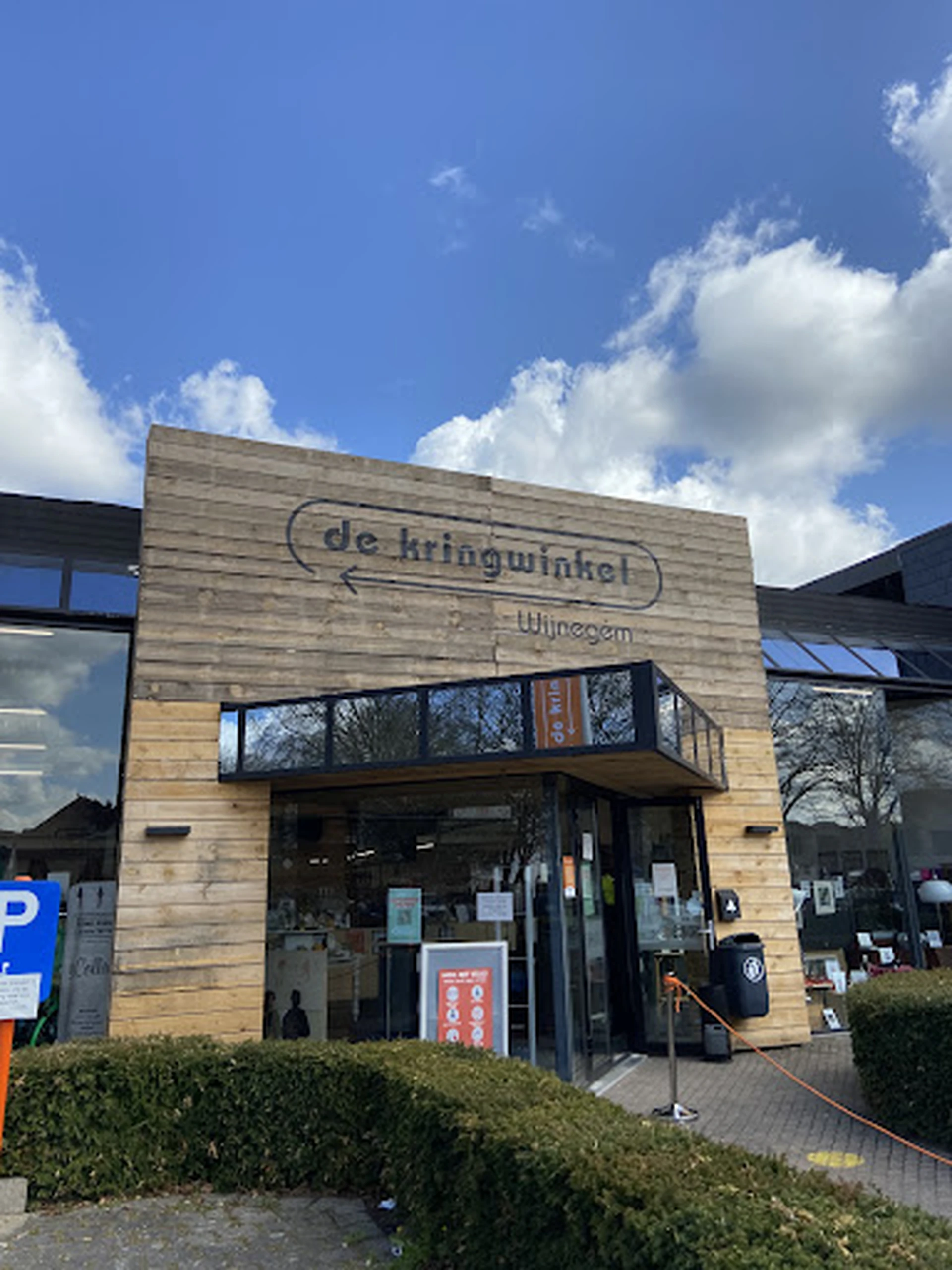 Houten gevel van kringloopwinkel met bord 'de kringwinkel' en naam 'Wijnegem', parkeerplaats zichtbaar