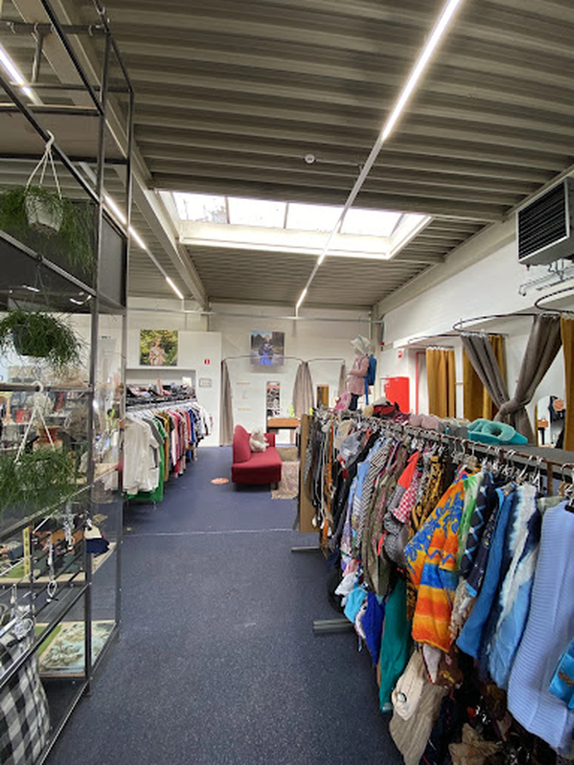 Kringloopwinkel met rekken vol kleding, groene planten, rode bankje en natuurlijke daglichtverlichting van bovenaf