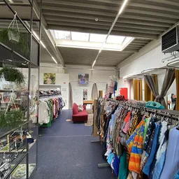 Kringloopwinkel met rekken vol kleding, groene planten, rode bankje en natuurlijke daglichtverlichting van bovenaf