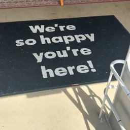 Zwart deurmat met witte tekst 'We're so happy you're here!'