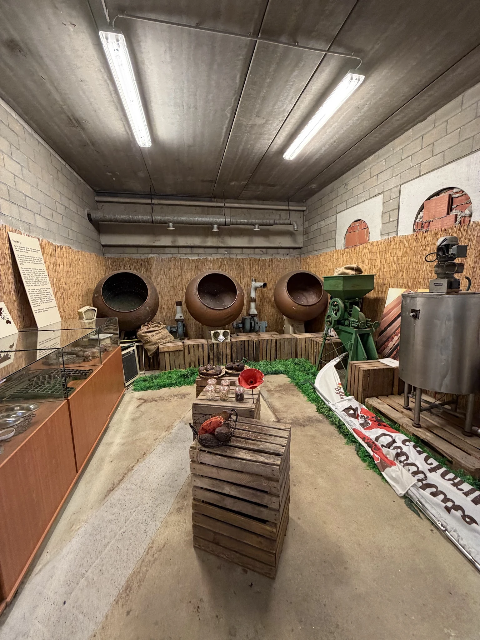 Museumachtige tentoonstelling met grote aardewerken potten, oude landbouwwerktuigen en houten kratten op een zandvloer
