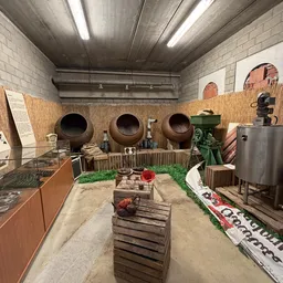 Museumachtige tentoonstelling met grote aardewerken potten, oude landbouwwerktuigen en houten kratten op een zandvloer
