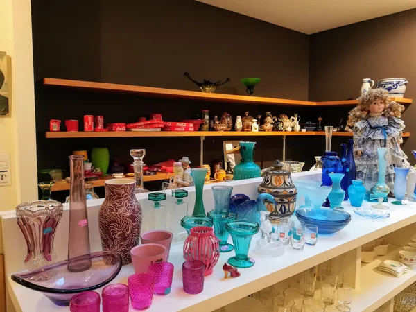 Uitstalling van gekleurde glazen vazen, kandelaars en decoratieve artikelen op witte toonbank met donkere muurplanken op acht