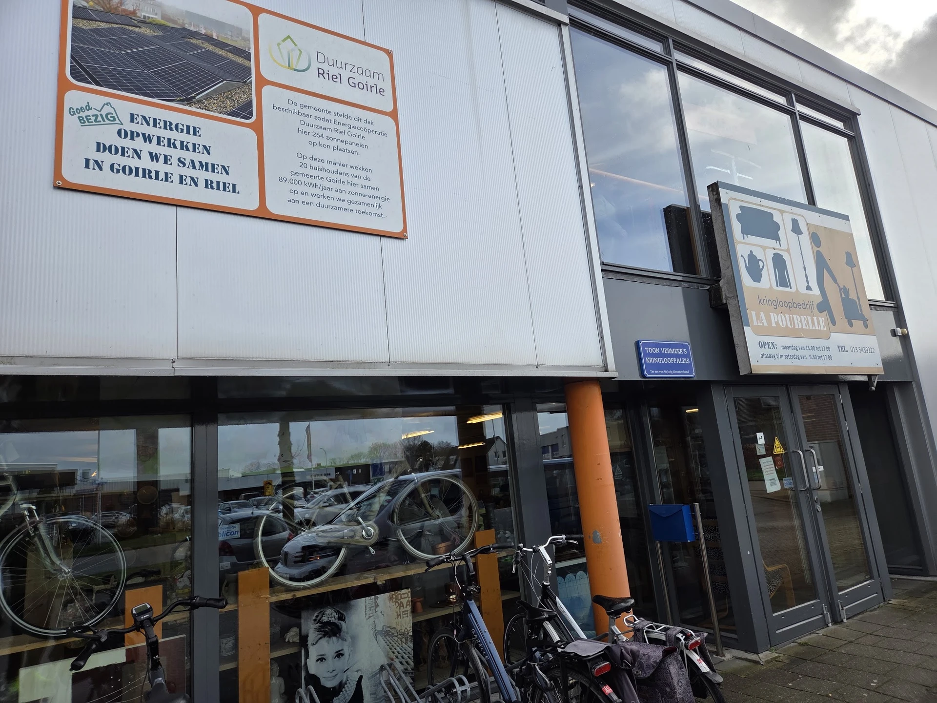 Voorgevel van kringloopwinkel met ramen, oranje accenten en fietsen in de etalage