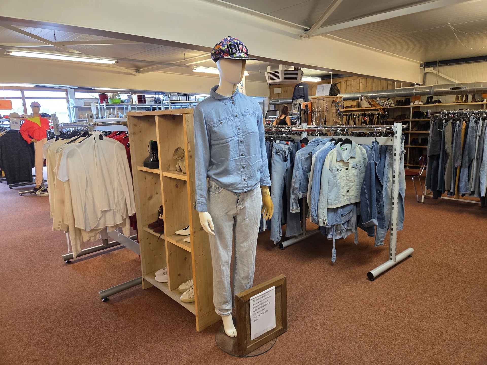 Etalage met mannequin in blauwe blouse en beige broek, omgeven door rekken met tweedehands kleding