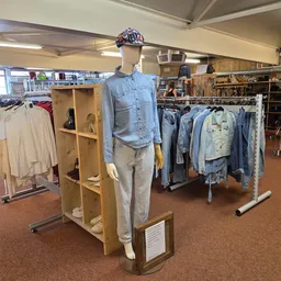 Etalage met mannequin in blauwe blouse en beige broek, omgeven door rekken met tweedehands kleding