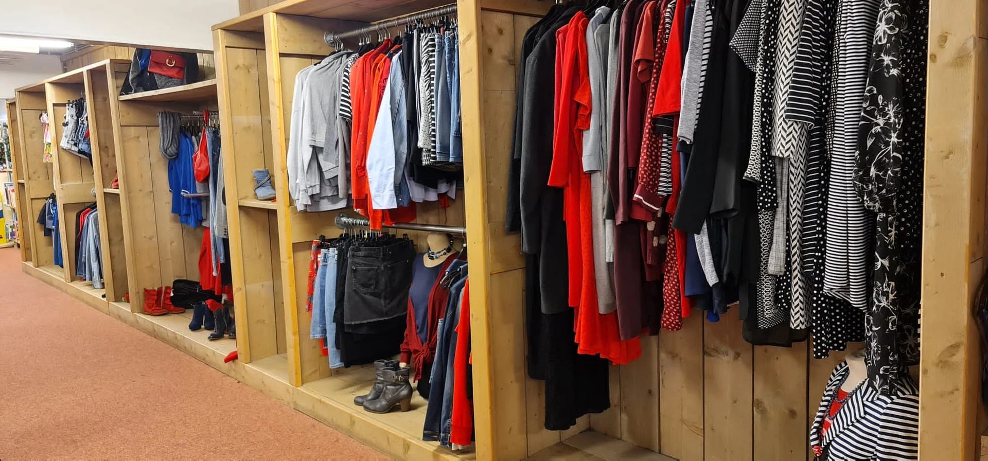 Houten stellingkast met tweedehands kleding in verschillende kleuren, waaronder rode, zwarte en grijze kledingstukken op hang