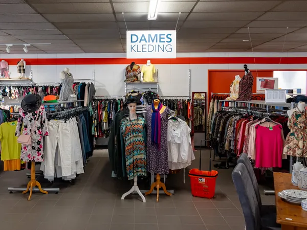 Winkelinterieur met rekken vol tweedehands dameskleding, witte en rode wanden, en een signage 'DAMES KLEDING' aan het plafond