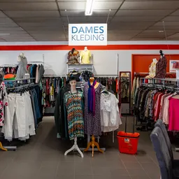 Winkelinterieur met rekken vol tweedehands dameskleding, witte en rode wanden, en een signage 'DAMES KLEDING' aan het plafond