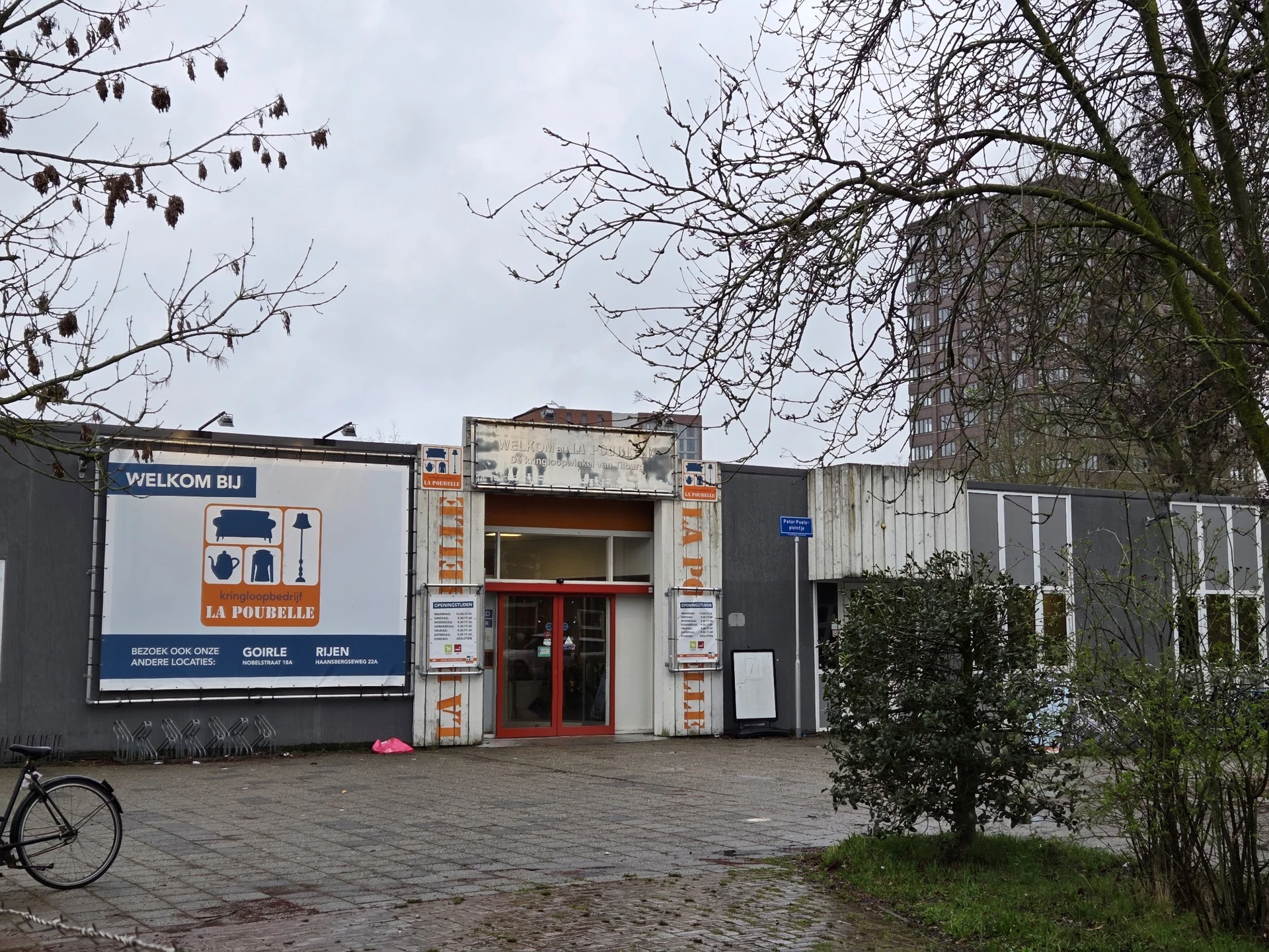 Gevel van kringloopwinkel La Poubelle met rode deur, welkomstbord en grijze aanbouw
