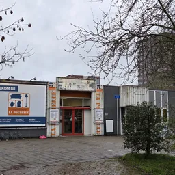 Gevel van kringloopwinkel La Poubelle met rode deur, welkomstbord en grijze aanbouw