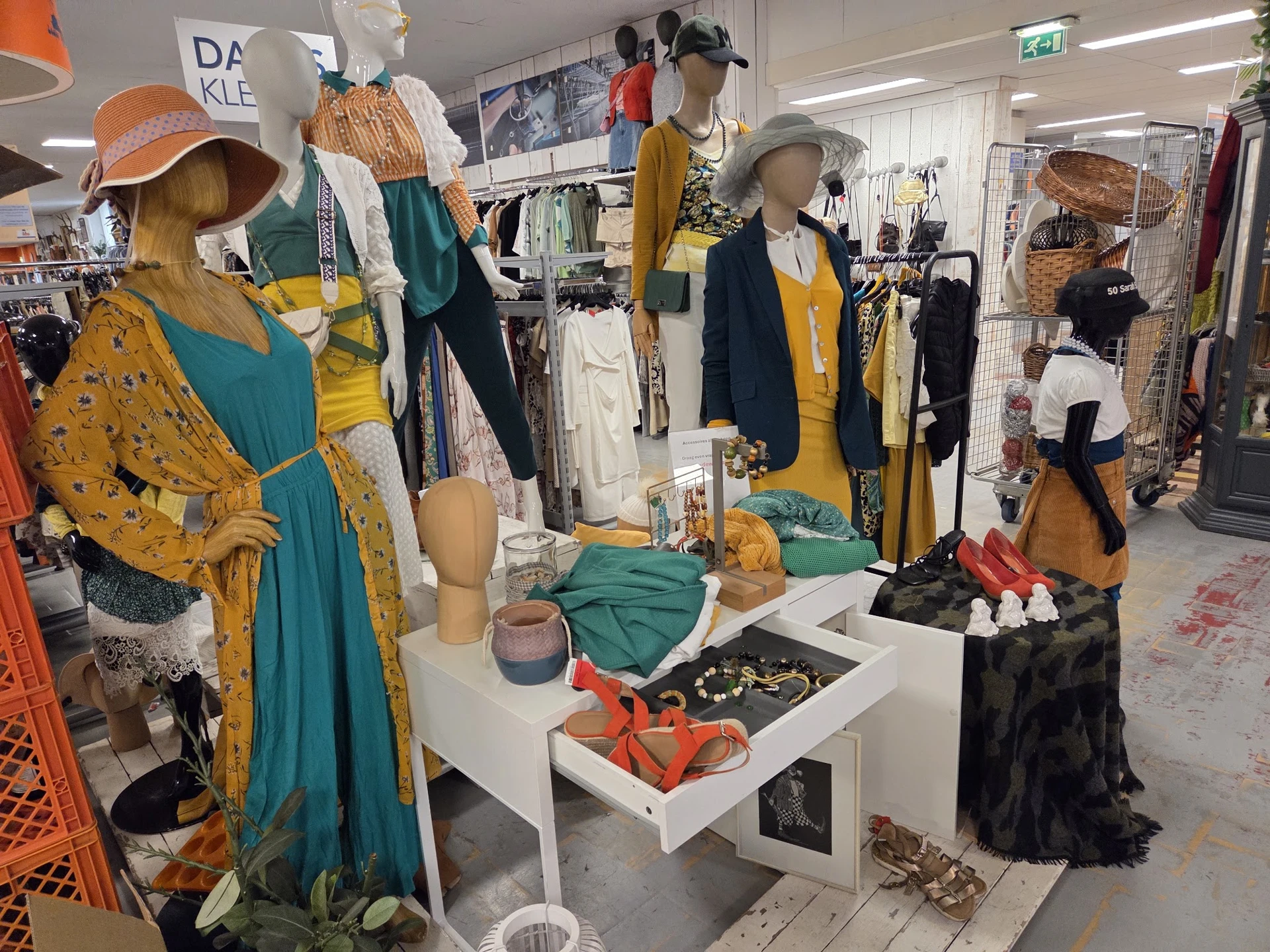 Etalage met mannequins gekleed in tweedehands kleding in geel, groen en blauw; accessoires en tassen op display