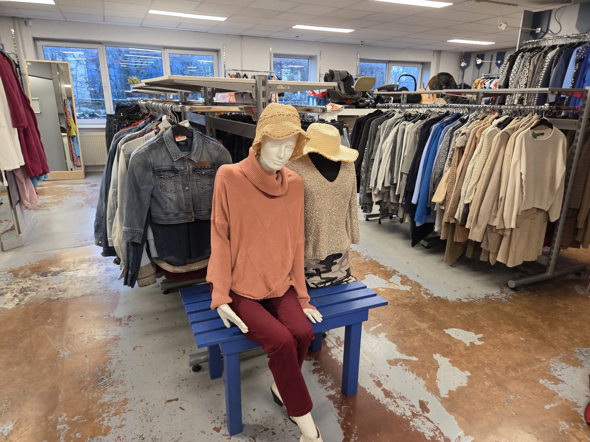Etalagepop in oranje trui en rode broek met gele pet, omgeven door rekken met tweedehands kleding