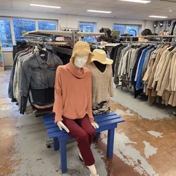 Etalagepop in oranje trui en rode broek met gele pet, omgeven door rekken met tweedehands kleding