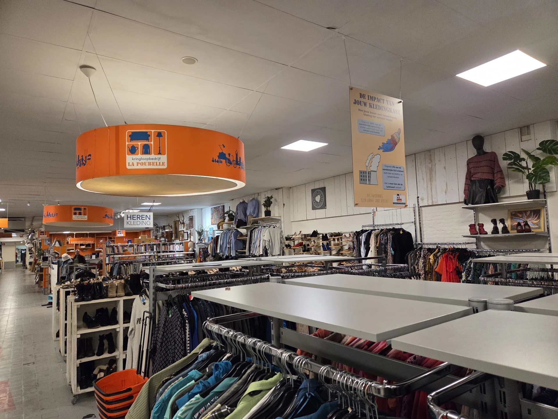 Kringloopwinkel met kledingrekken, tafels met kledingstukken en een opvallende oranje hanglamp.