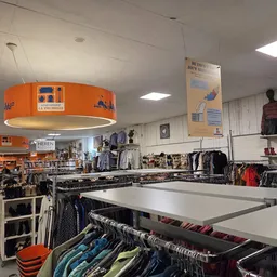 Kringloopwinkel met kledingrekken, tafels met kledingstukken en een opvallende oranje hanglamp.