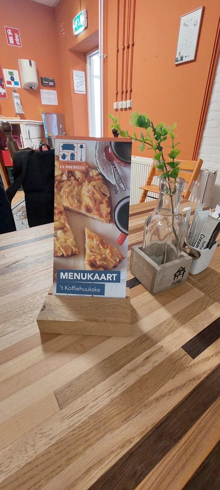 Menukaart 'La Poubelle 't Koffihuukske' met afbeelding van gebak, op tafel in de winkel