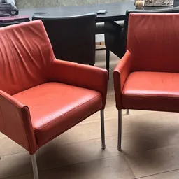Twee rode/koraalkleurige fauteuils met armleuningen en metalen poten naast elkaar op een betonnen vloer