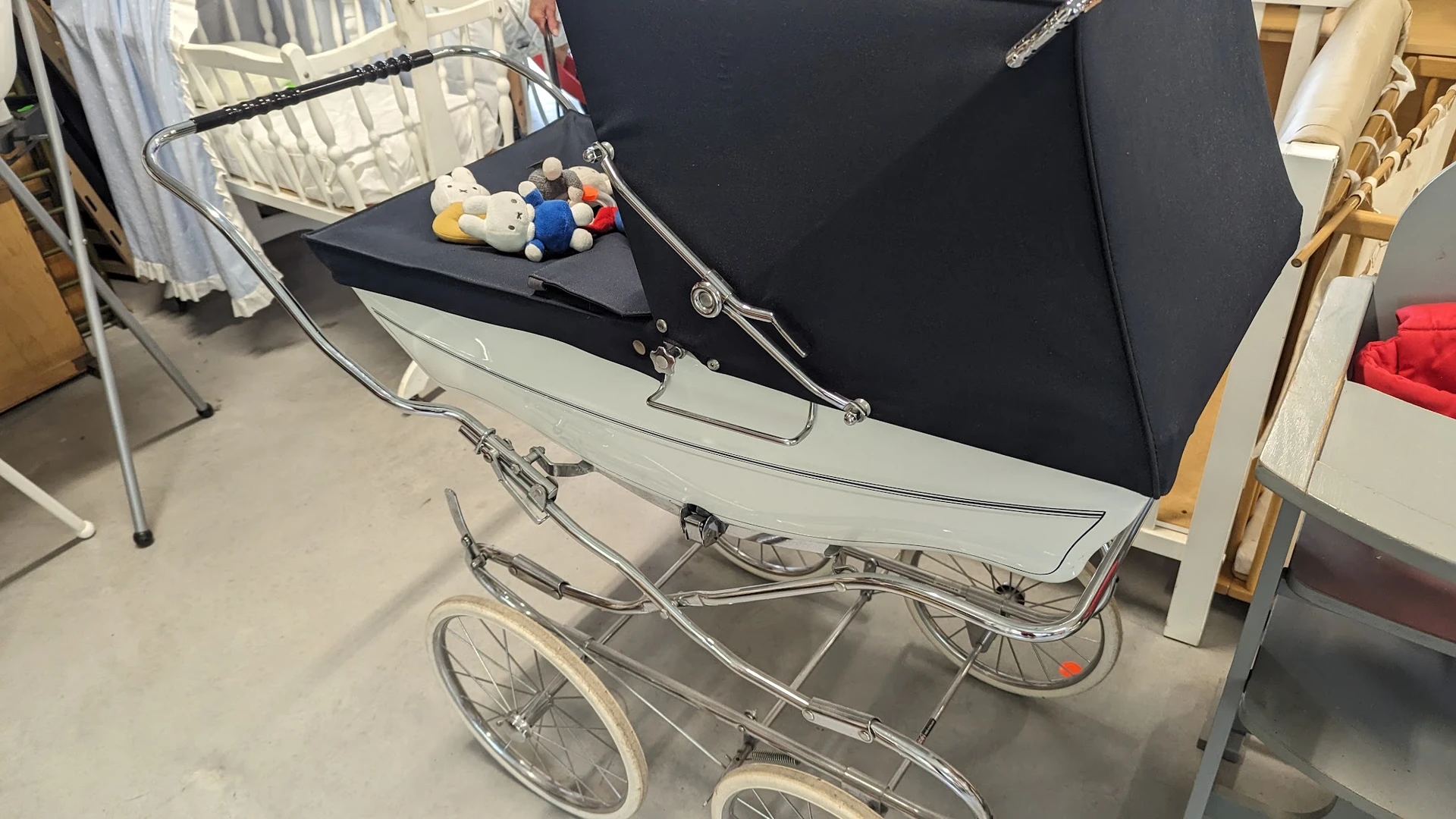 Vintage zilvergrijze kinderwagen met zwart zonnedak en speelgoedset bovenop, in een kringloopwinkel