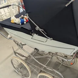 Vintage zilvergrijze kinderwagen met zwart zonnedak en speelgoedset bovenop, in een kringloopwinkel
