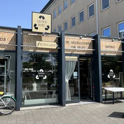 Gevel van kringloopwinkel met zwarte puien, grote ramen en uithangborden met winkelnaam, witte fiets ervoor
