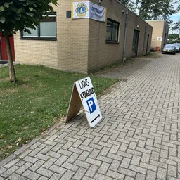 Parkeerplaats en grasveld voor een bakstenen gebouw met een A-bord met parkeerlogo