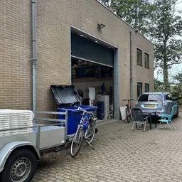 Voorkant van een bakstenen pand met open garagedeur, daarbuiten een blauwe bakfiets, auto en kampeerstoelen op het terras
