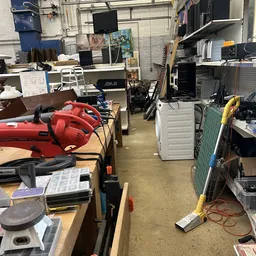Rommelige werkplaats of opslagruimte met rode elektrogereedschappen, witte apparaten en diverse spullen op houten tafels en s