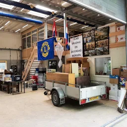 Werkplaats en opslagruimte van kringloopwinkel met stellingen, werkbanken, aanhanger en Lions-vlag