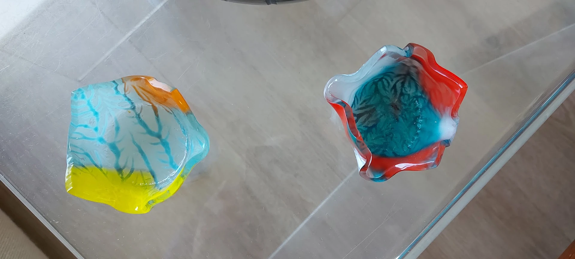 Twee gekleurde glazen decoratieve voorwerpen met blauw, rood, geel en oranje tinten op een witte oppervlak