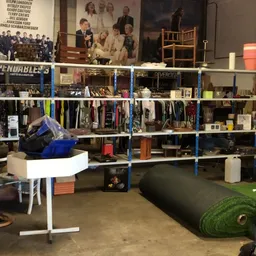 Winkelvloer met blauwe stellingen vol kleding, boeken en huishoudspullen, kunstgras en diverse meubelstukken