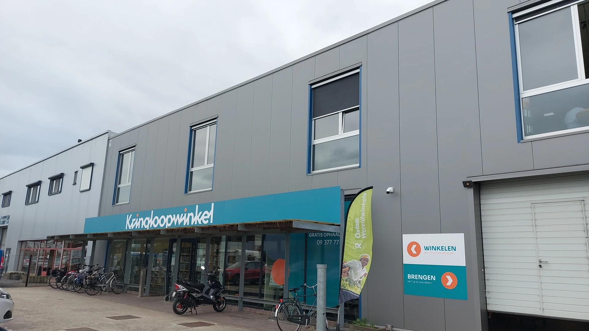 Grijze gevel van kringloopwinkel met turquoise uitbouw en winkelnaam, ramen en ingang met fietsen