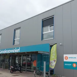 Grijze gevel van kringloopwinkel met turquoise uitbouw en winkelnaam, ramen en ingang met fietsen
