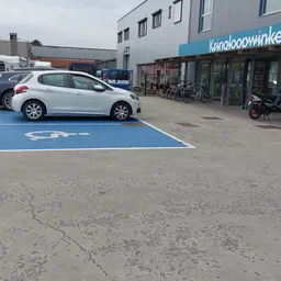 Witte auto geparkeerd op elektrisch voertuig laadplek voor een kringloopwinkel met winkelpui op achtergrond