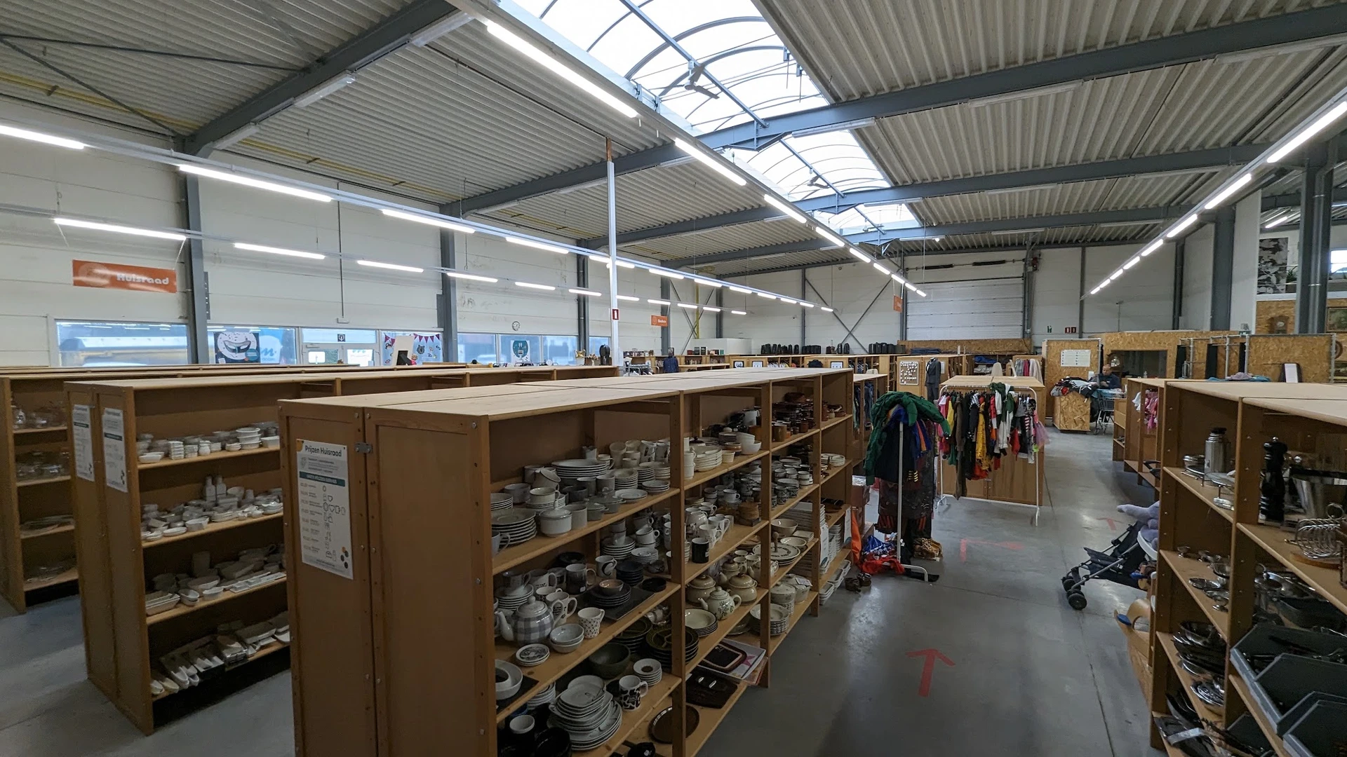 Winkelvloer met houten stellingen vol servies en huishoudartikelen, kledingrekken op de achtergrond