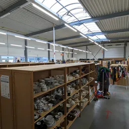 Winkelvloer met houten stellingen vol servies en huishoudartikelen, kledingrekken op de achtergrond