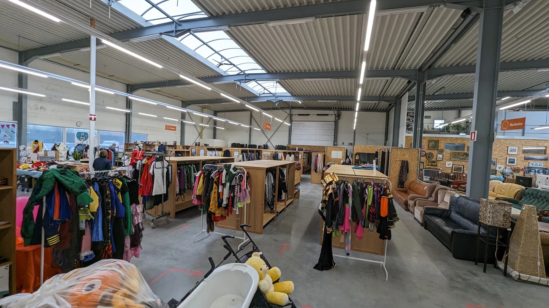 Ruime winkelvloer met houten stellingen vol tweedehands kleding, meubelstukken en huishoudspullen onder industrieel plafond