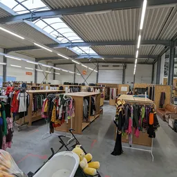 Ruime winkelvloer met houten stellingen vol tweedehands kleding, meubelstukken en huishoudspullen onder industrieel plafond