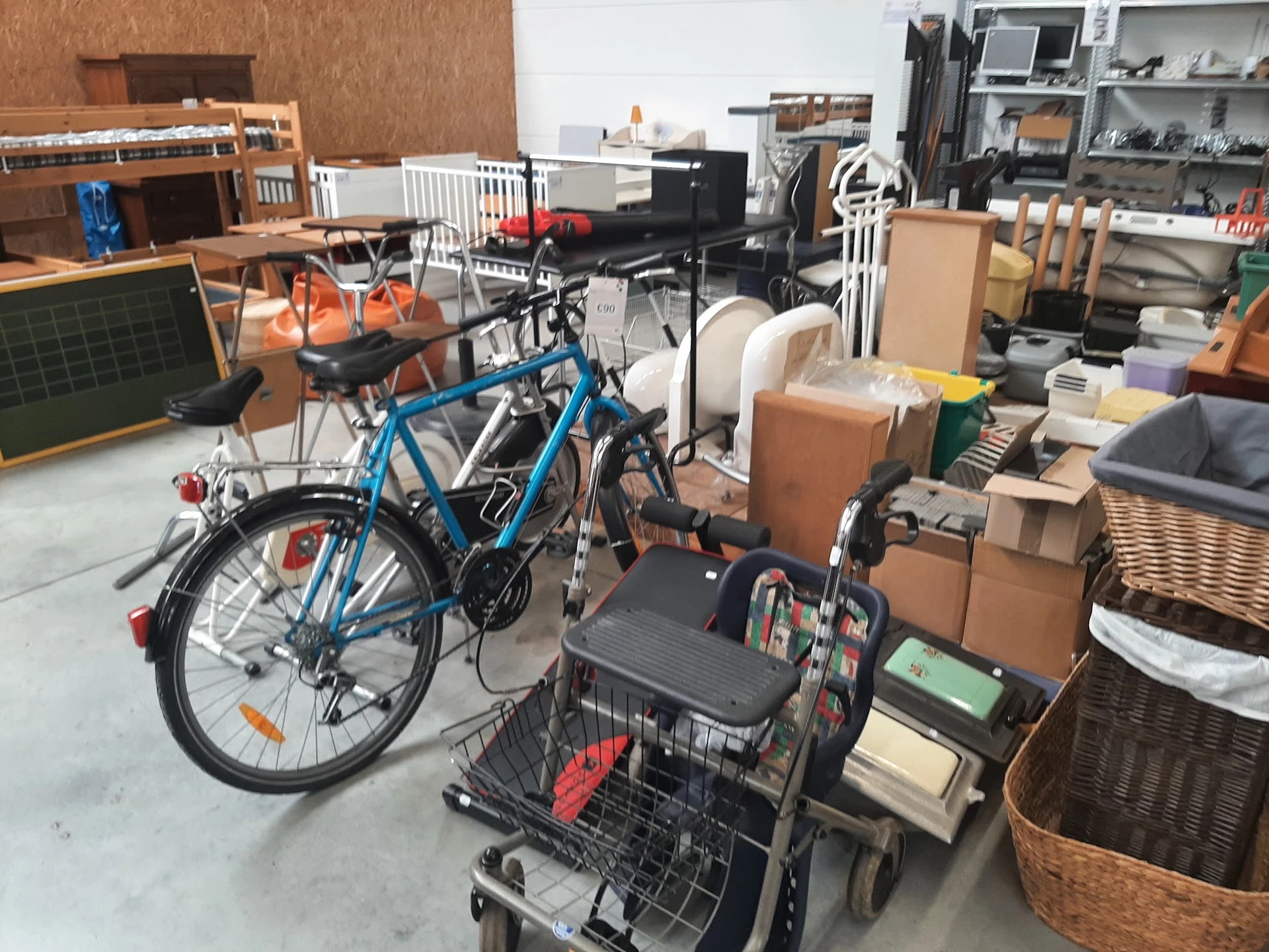 Binnenaanzicht van kringloopwinkel met blauwe fiets, winkelwagen en dozen met huishoudelijke spullen