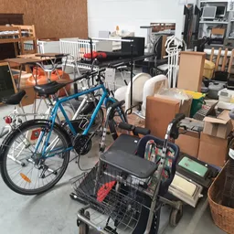 Binnenaanzicht van kringloopwinkel met blauwe fiets, winkelwagen en dozen met huishoudelijke spullen