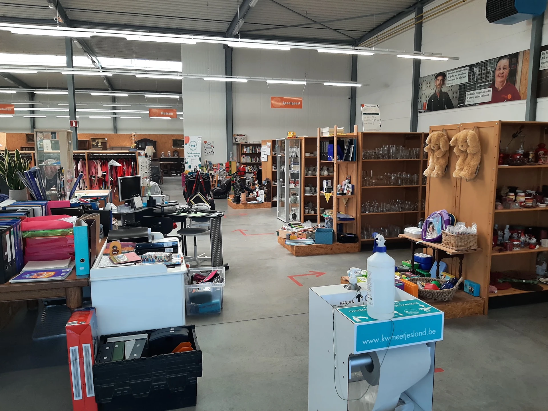 Ruime winkelruimte met diverse stellingen, kleding, boeken en huishoudartikelen verspreid over de vloer