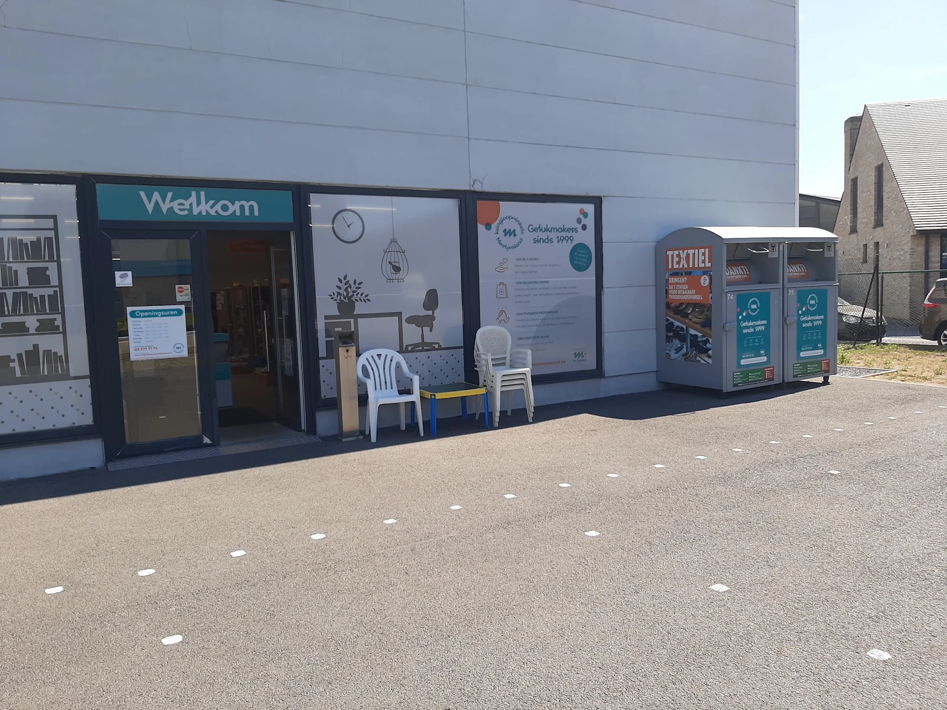 Voorkant van kringloopwinkel met Welkom-ingang, uitstalramen, informatieborden en textielcontainers