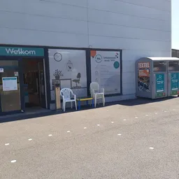 Voorkant van kringloopwinkel met Welkom-ingang, uitstalramen, informatieborden en textielcontainers