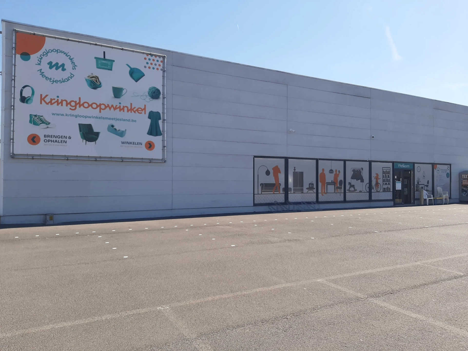 Grijze gevel van kringloopwinkel met logo en uitstalbakken, parkeerterrein ervoor