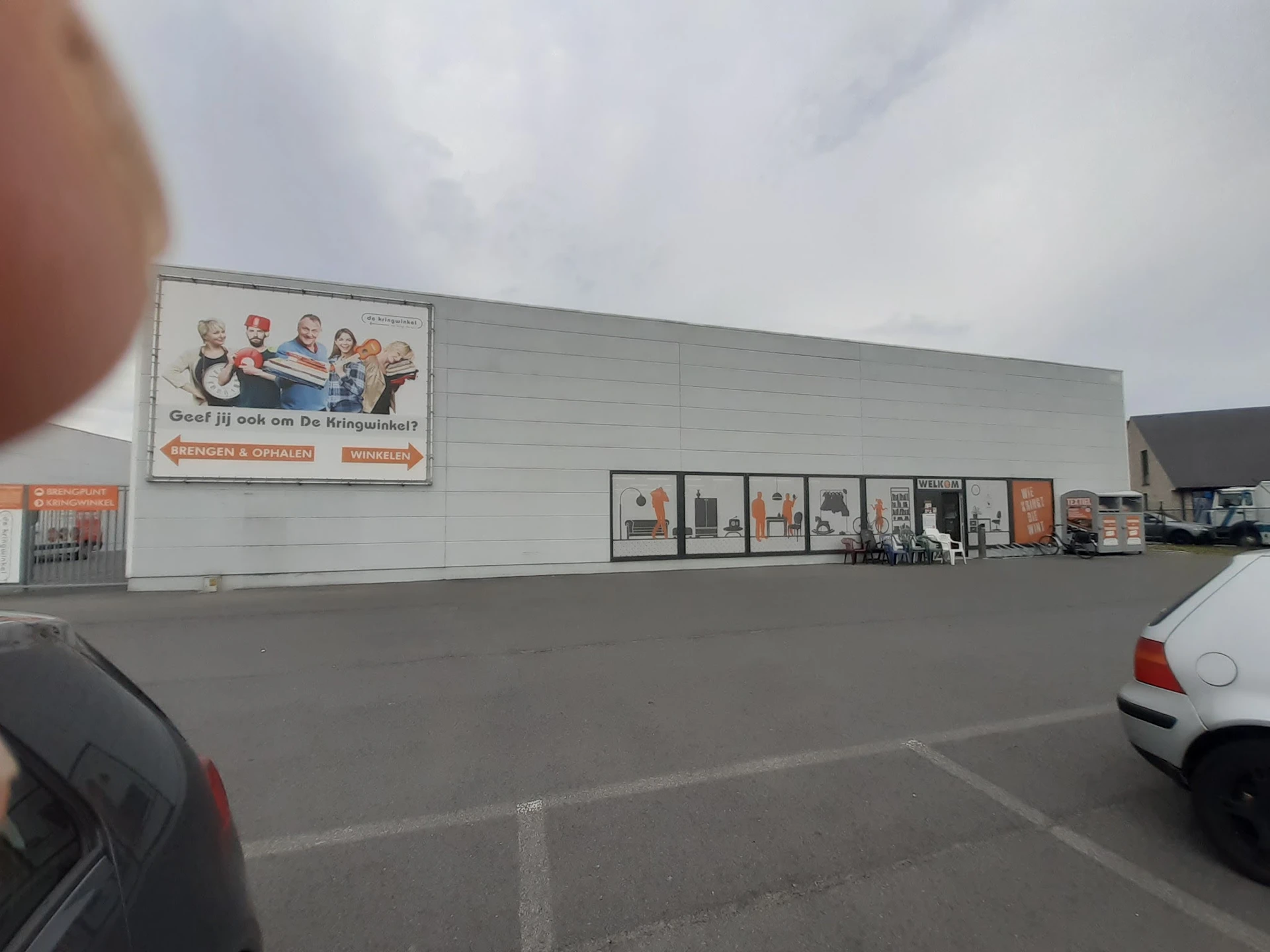 Voorgevel van kringloopwinkel met witte muur, bewegwijzering en parkeerplaats met auto's
