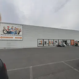 Voorgevel van kringloopwinkel met witte muur, bewegwijzering en parkeerplaats met auto's